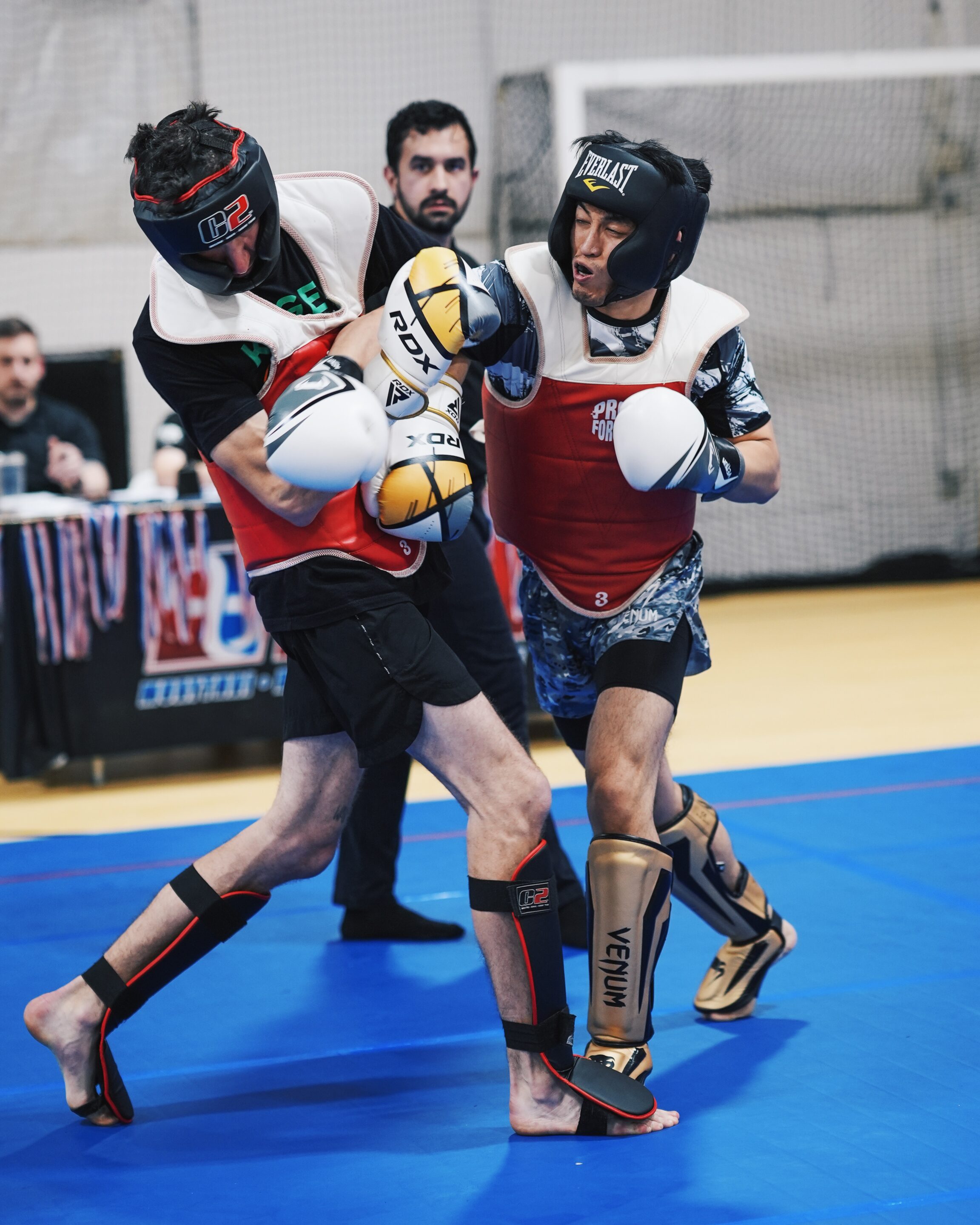 Two male fighters in headgear and padded vests clash while a referee watches in the background on a blue-surface gym floor.