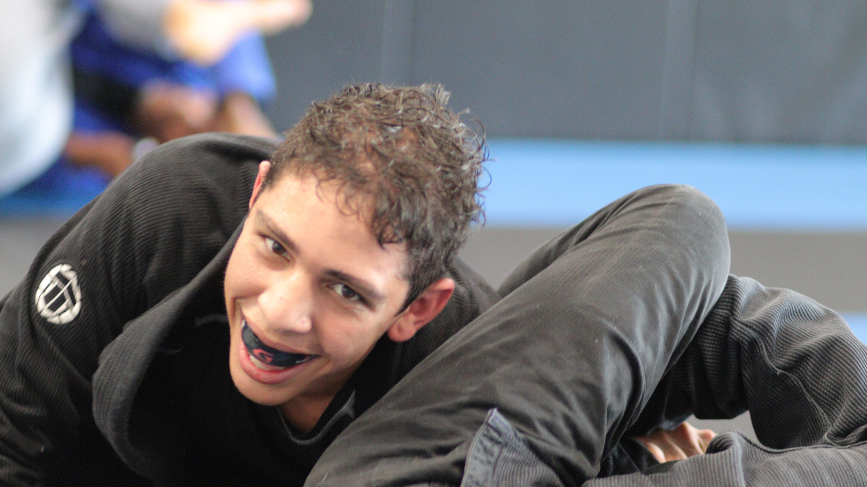 Young man in a black gi smiling while grappling on a gym mat.